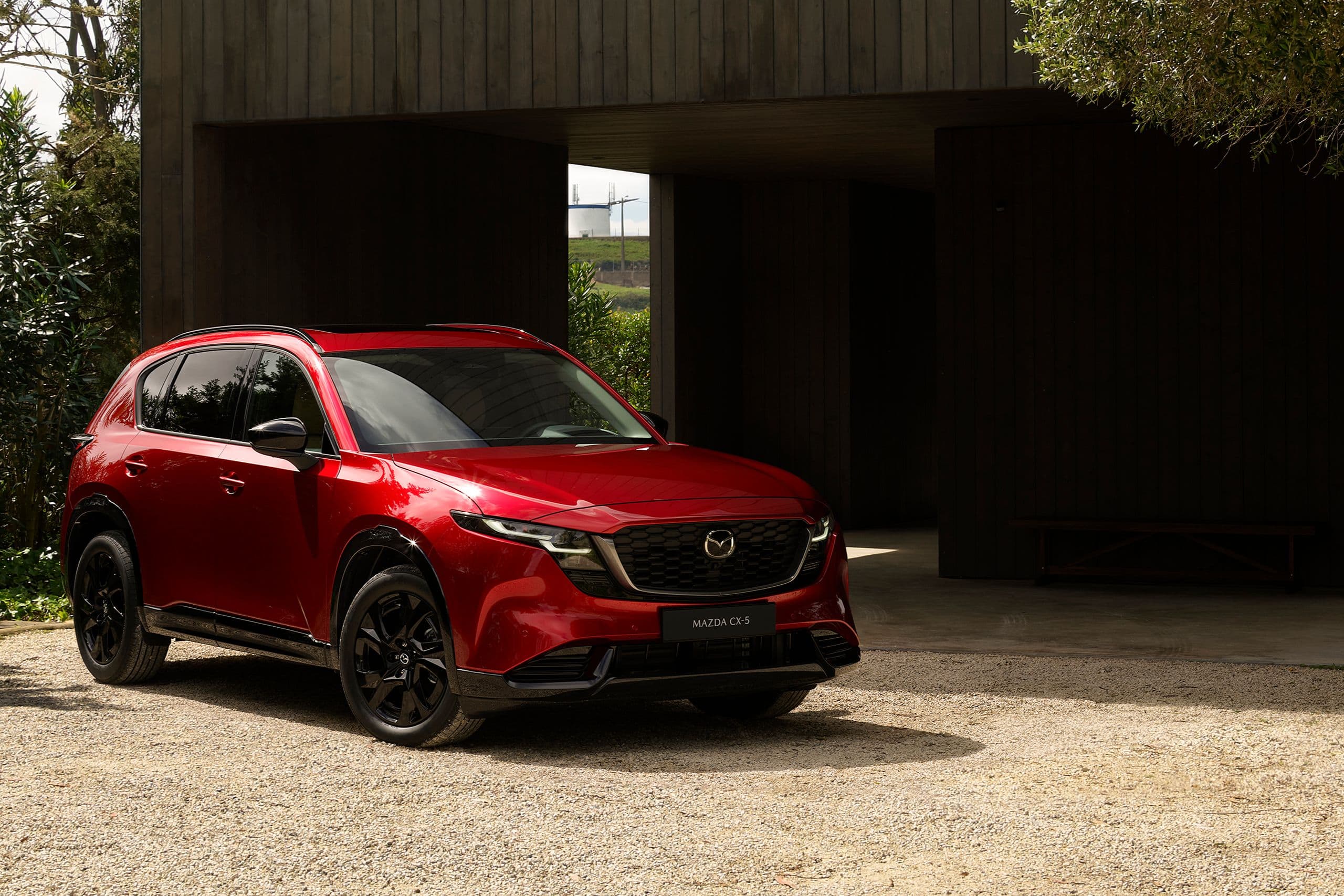 Mazda CX-5 2025 in Soul Red Crystal geparkt vor einem modernen Holzhaus mit natürlicher Umgebung – elegantes Exterieurdesign im Sonnenlicht.